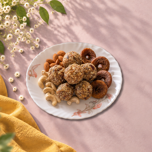 VERU Foods Mewa Laddoo plate styled on a soft pastel pink textured background with almonds, cashews, dried figs, flowers and warm mustard fabric.
